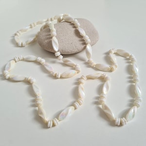 Könnte beinhalten: Eine lange weiße Halskette aus schimmernden ovalen Perlen. Die Kette ist auf einer weißen Schnur aufgefädelt und liegt auf einer weißen Oberfläche.