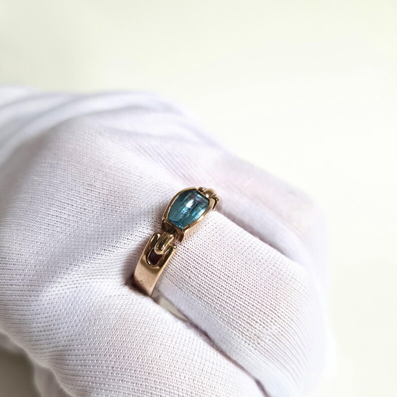 May include: A gold-toned ring featuring a rectangular, light blue gemstone. The ring has a unique design with curved accents on the band and around the stone. The gemstone is set in a bezel setting, enhancing its color and shape. A piece of jewelry.