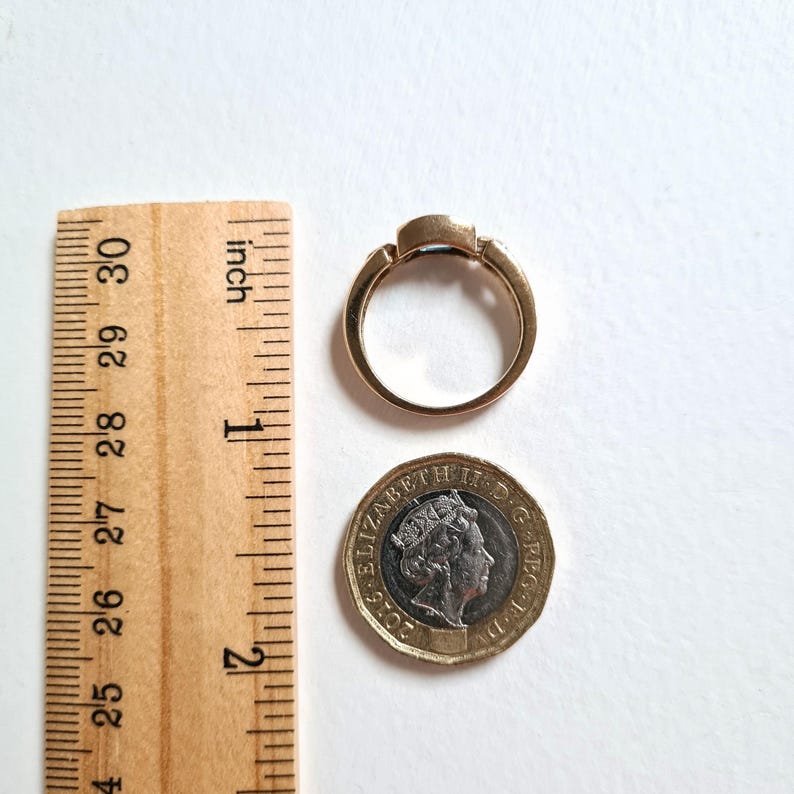 May include: A gold ring with a rectangular gemstone, next to a wooden ruler and a coin. The ring's band is a warm gold color, and the gemstone is a darker color. The ruler is marked in inches.