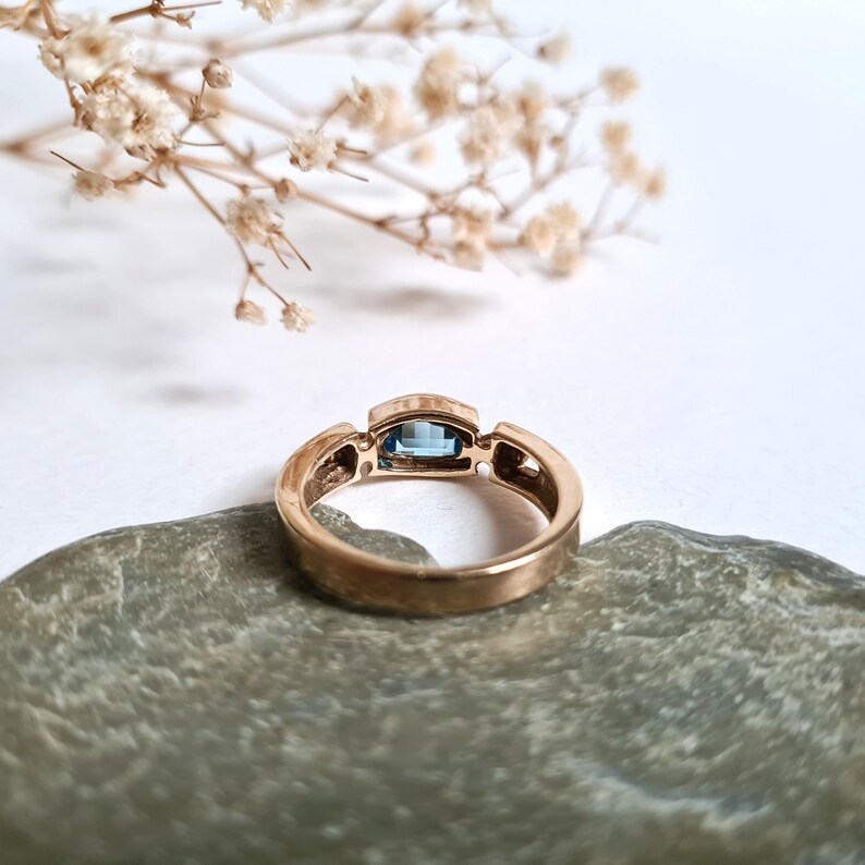 May include: A gold ring with a rectangular, light blue gemstone. The ring is set on a gray stone, with dried flowers in the background. The ring's band has a textured design.