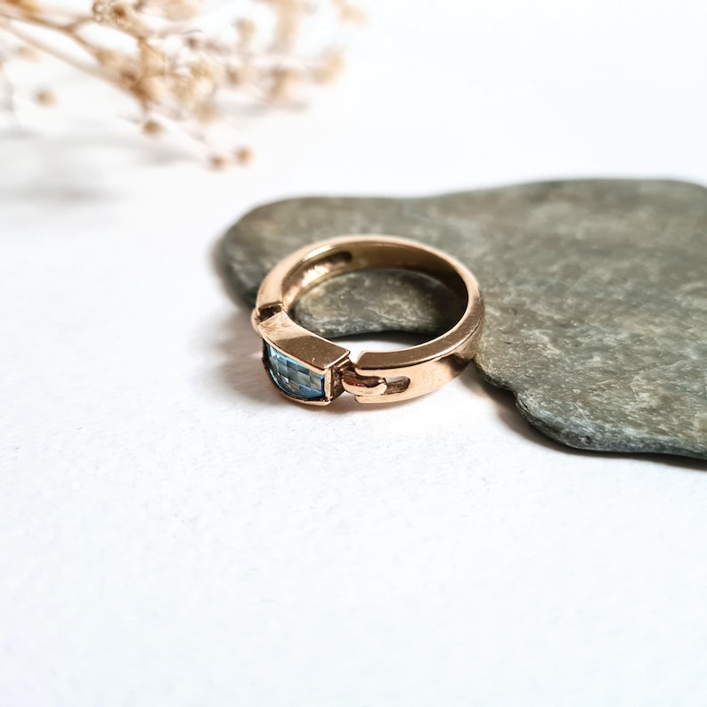 May include: A gold-toned ring with a rectangular, light blue gemstone. The ring is set on a gray stone, with a white background. The ring's design features a unique clasp detail on the sides of the gemstone setting. A sprig of dried flowers is in the background.