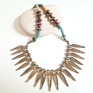 May include: A gold necklace with a long, layered chain and a pendant of multiple gold leaves. The chain is made of a mix of brown, red, and blue cords with metal accents.
