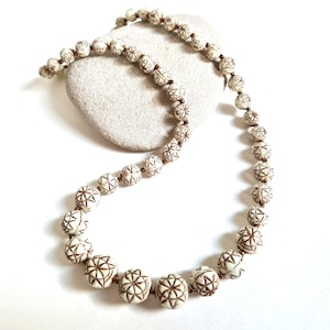 Puede incluir: Un collar de cuentas color crema con patrones geométricos marrones. El collar está colocado sobre una piedra clara sobre un fondo blanco. Las cuentas son esféricas y están ensartadas con un cordón marrón.