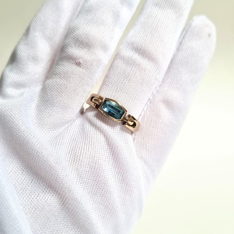 May include: A gold ring featuring a rectangular, light blue gemstone. The ring has a simple band with decorative elements on either side of the gemstone. The ring is displayed on a white surface.