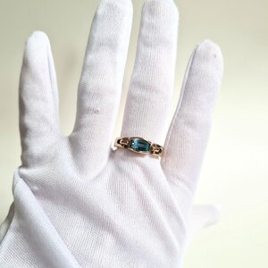 May include: A gold ring with a rectangular, light blue gemstone. The ring is set in a gold band with decorative accents on either side of the gemstone. The ring is displayed on a white glove.