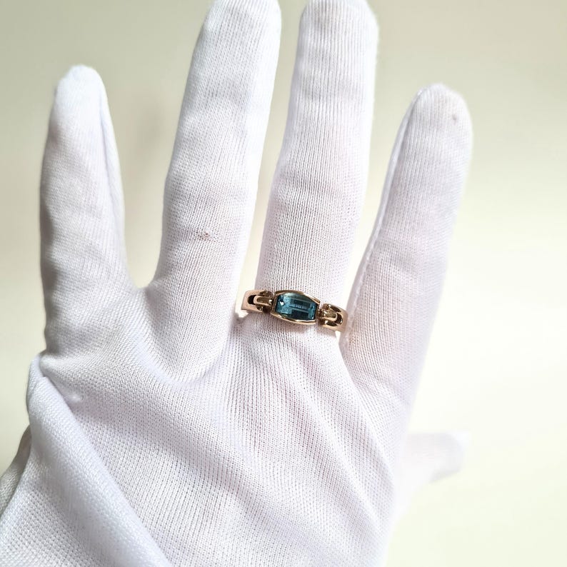 May include: A gold ring with a rectangular, light blue gemstone. The ring is set in a gold band with decorative accents on either side of the gemstone. The ring is displayed on a white glove.