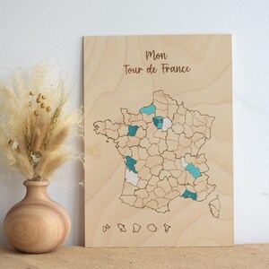 May include: Wooden map of France with teal and white accents marking locations. The text "Mon Tour de France" is at the top. A wooden vase with dried flowers is to the left of the map.