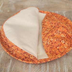 May include: A round, orange and brown fabric pot holder with a white cotton lining. The pot holder is folded in half, showing the lining.