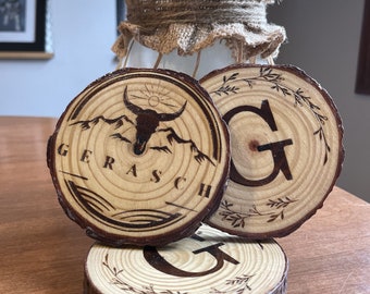 May include: Three wooden coasters with a rustic design. One coaster features a bull skull and mountain graphic with the word "GERASCH". Another has the letter "G" with a leaf design. The coasters are stacked on a wooden surface.