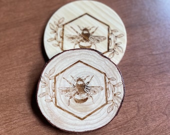 May include: Two wooden coasters with a bee design. The bee is inside a hexagon with leaves around it. The coasters are made of natural wood and have a rustic look.