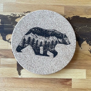 May include: A round cork coaster featuring a black and white illustration of a bear walking through a forest. The bear is silhouetted against a mountain range with trees.