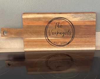 May include: A wooden cutting board with a handle. The board is engraved with a circle containing the text "The Vorpagels EST. 2001".
