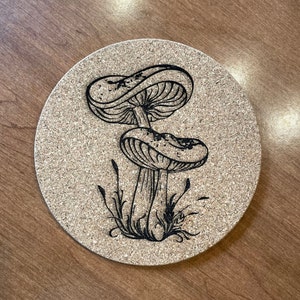 May include: A cork coaster with a black and white illustration of two mushrooms with a stem and a cap. The mushrooms are surrounded by grass.