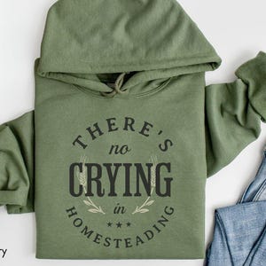 May include: A military green hoodie with the text "THERE'S NO CRYING in HOMESTEADING" in a circular design. The hoodie has a drawstring hood and long sleeves. A pair of blue jeans is visible in the background.