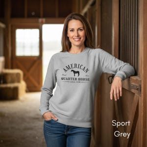 K&ouml;nnte beinhalten: Ein hellgraues Sweatshirt mit dem Text "American Quarter Horse Since 1940" in Schwarz. Das Sweatshirt zeigt eine Grafik eines Pferdes. Das Model tr&auml;gt eine blaue Jeans. Das Sweatshirt ist mit "Sport Grey" beschriftet.