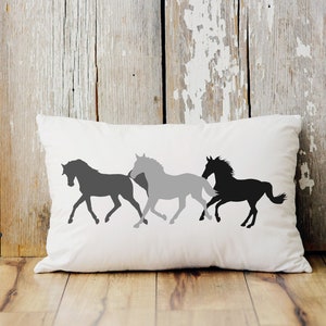 May include: White rectangular pillow featuring three running horse silhouettes in shades of gray and black. The pillow is placed on a wooden surface with a distressed wood background.