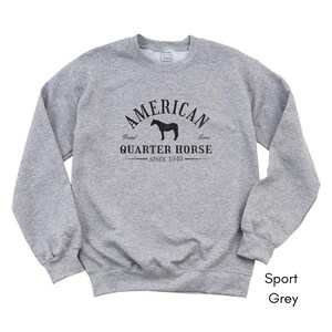 K&ouml;nnte beinhalten: Ein hellgraues Sweatshirt mit dem schwarzen Text "AMERICAN QUARTER HORSE SINCE 1940". Das Design beinhaltet eine Pferdesilhouette. Das Sweatshirt ist mit "Sport Grey" gekennzeichnet.