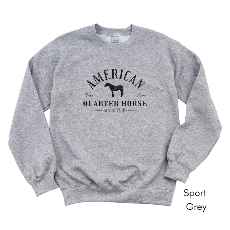 K&ouml;nnte beinhalten: Ein hellgraues Sweatshirt mit dem schwarzen Text "AMERICAN QUARTER HORSE SINCE 1940". Das Design beinhaltet eine Pferdesilhouette. Das Sweatshirt ist mit "Sport Grey" gekennzeichnet.