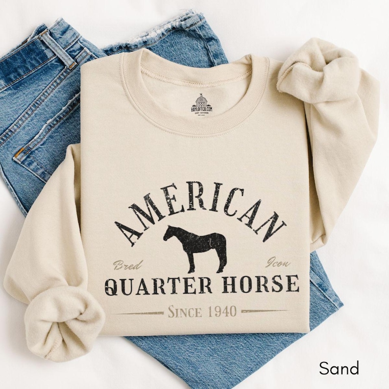 K&ouml;nnte beinhalten: Ein sandfarbenes Sweatshirt mit dem schwarzen Text "AMERICAN QUARTER HORSE" und einer Pferdesilhouette. Das Sweatshirt enth&auml;lt den Text "Since 1940" und liegt auf einer Jeans.
