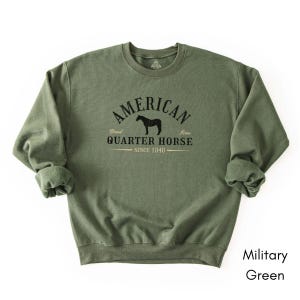 K&ouml;nnte beinhalten: Milit&auml;rgr&uuml;nes Sweatshirt mit dem schwarzen Text "AMERICAN QUARTER HORSE SINCE 1940" und einer Pferdesilhouette. Das Sweatshirt hat einen Rundhalsausschnitt.