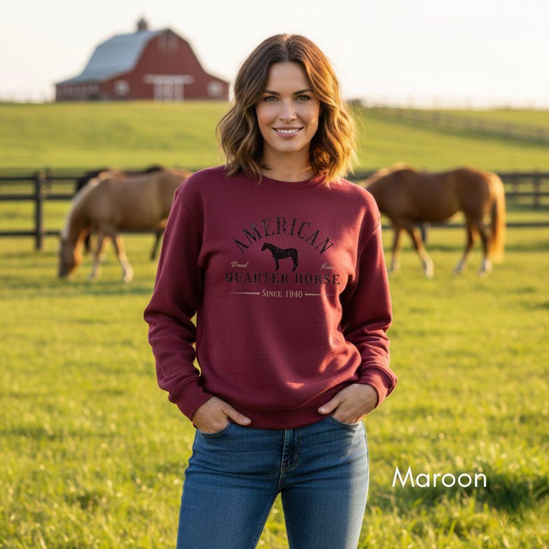 K&ouml;nnte beinhalten: Burgunderfarbenes Sweatshirt mit dem Text "AMERICAN QUARTER HORSE SINCE 1940" und einer Pferdesilhouette. Das Sweatshirt wird mit einer blauen Jeans kombiniert. Der Hintergrund zeigt eine gr&uuml;ne Wiese, Pferde, eine rote Scheune und einen Holzzaun.