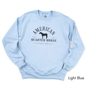 K&ouml;nnte beinhalten: Hellblaues Sweatshirt mit dem Text "AMERICAN QUARTER HORSE" und einer Pferdesilhouette. Der Text "Since 1940" ist ebenfalls auf dem Sweatshirt aufgedruckt. Das Sweatshirt hat einen Rundhalsausschnitt.