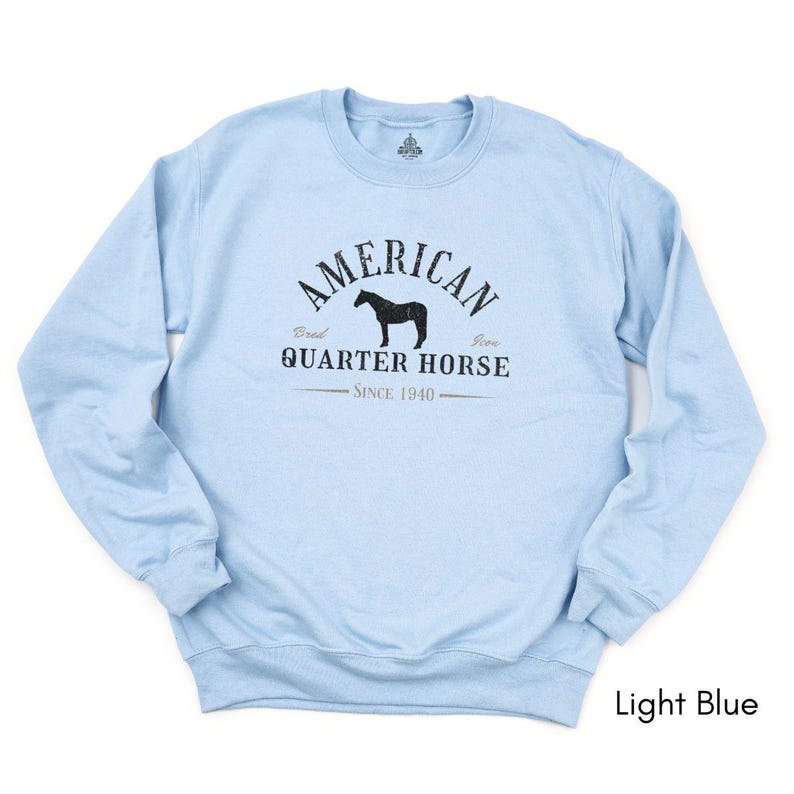 K&ouml;nnte beinhalten: Hellblaues Sweatshirt mit dem Text "AMERICAN QUARTER HORSE" und einer Pferdesilhouette. Der Text "Since 1940" ist ebenfalls auf dem Sweatshirt aufgedruckt. Das Sweatshirt hat einen Rundhalsausschnitt.