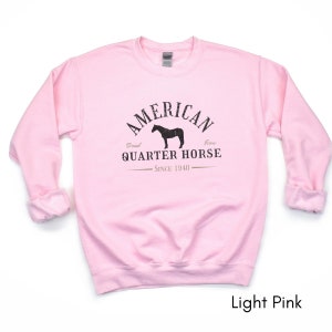 K&ouml;nnte beinhalten: Hellrosa Sweatshirt mit dem Text "AMERICAN QUARTER HORSE" in verblichener schwarzer Schrift, zusammen mit einer Pferdesilhouette. Der Text "Bred Icon Since 1940" ist ebenfalls enthalten. Das Sweatshirt hat lange &Auml;rmel und einen Rundhalsausschnitt.