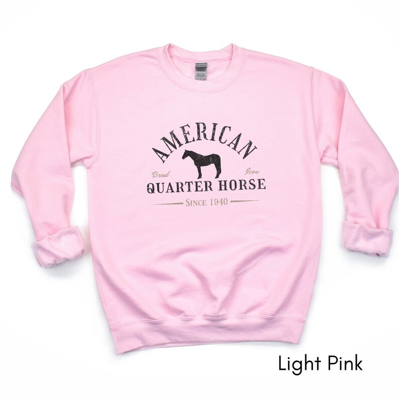 K&ouml;nnte beinhalten: Hellrosa Sweatshirt mit dem Text "AMERICAN QUARTER HORSE" in verblichener schwarzer Schrift, zusammen mit einer Pferdesilhouette. Der Text "Bred Icon Since 1940" ist ebenfalls enthalten. Das Sweatshirt hat lange &Auml;rmel und einen Rundhalsausschnitt.
