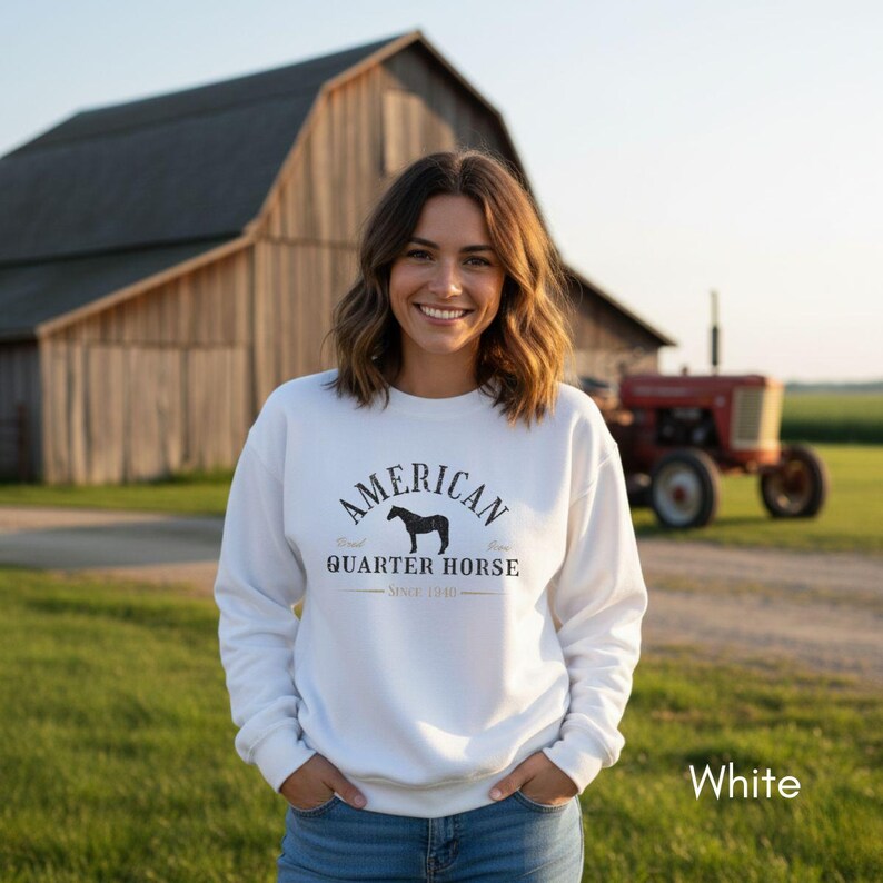 K&ouml;nnte beinhalten: Wei&szlig;es Sweatshirt mit dem Text "AMERICAN QUARTER HORSE SINCE 1940" in Schwarz. Es zeigt eine Pferdesilhouette. Im Hintergrund sind eine Scheune, ein Traktor und ein gr&uuml;nes Feld zu sehen.