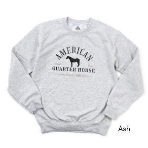 K&ouml;nnte beinhalten: Hellgraues Sweatshirt mit dem schwarzen Schriftzug "AMERICAN QUARTER HORSE SINCE 1940" und einer Pferdesilhouette. Das Sweatshirt hat einen Rundhalsausschnitt und lange &Auml;rmel. Die Farbe ist "Ash".