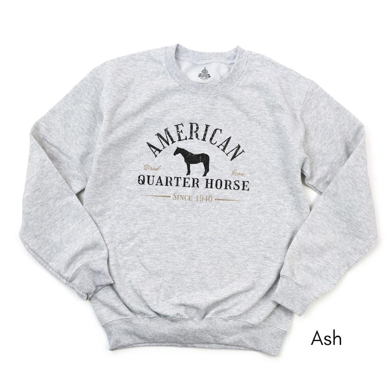 K&ouml;nnte beinhalten: Hellgraues Sweatshirt mit dem schwarzen Schriftzug "AMERICAN QUARTER HORSE SINCE 1940" und einer Pferdesilhouette. Das Sweatshirt hat einen Rundhalsausschnitt und lange &Auml;rmel. Die Farbe ist "Ash".