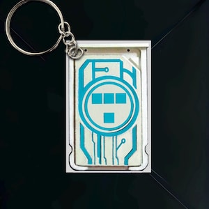 May include: A rectangular keychain with a silver keyring. The keychain features a white background with a blue circuit board design. The design includes a circle with three squares and a single square. The text "EPIC NERDZ NOVELTY CO." is at the bottom.