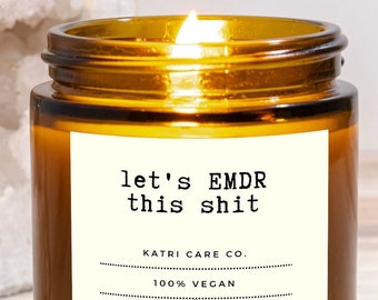 May include: A lit candle in an amber glass jar with a white label that reads let's EMDR this shit, KATRI CARE CO., 100% VEGAN, 20 HOURS, 4 OZ, and COCONUT & SOY CANDLE. The candle is positioned in front of a large, rough, white crystalline rock.