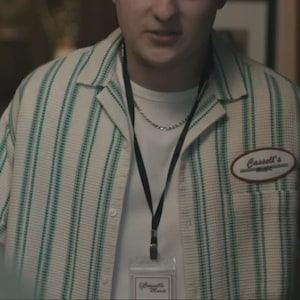 May include: A short-sleeved button-down shirt with a white and green vertical striped pattern, featuring a camp collar and an oval patch on the chest that reads Cassell's Music. A white undershirt, a silver chain, and a black lanyard are worn.