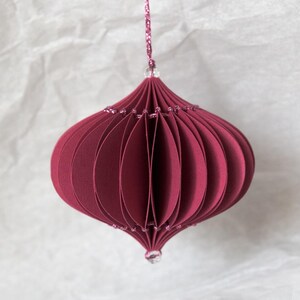 May include: A burgundy paper ornament with a layered, rounded design. The ornament is suspended by a glittery pink string and adorned with small, sparkling beads. The decorative piece is suitable for holiday or seasonal decor.