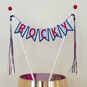 May include: A cake topper with the name "ROCKY" spelled out on a banner. The banner has a blue and white gingham pattern with red letters and is strung between two white poles topped with red spheres. Red, white, and blue ribbons hang from the poles.