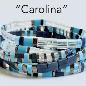 May include: A stack of blue, white, and silver beaded bracelets. The bracelets are stacked on top of each other and have a variety of different patterns. The text "Carolina" is written above the bracelets.