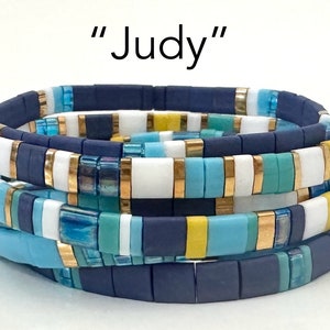 May include: A stack of four bracelets made of colorful ceramic tiles. The bracelets are in shades of blue, yellow, white, and green. The tiles are arranged in a repeating pattern. The bracelets are stacked on top of each other, with the top bracelet being the largest. The bracelets are called "Judy".