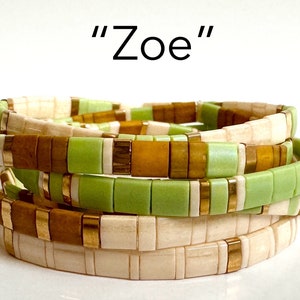May include: Stack of four tile bracelets in shades of cream, brown, and green with gold-colored accents. The word "Zoe" is written in black above the bracelets. The bracelets are stacked on top of each other.