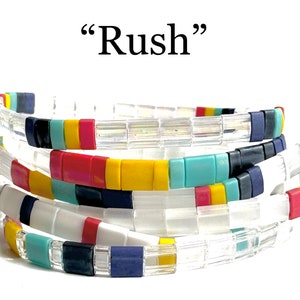 May include: Multicolored beaded bracelets stacked on top of each other. The bracelets are made of small, square beads in various colors, including white, red, yellow, blue, and turquoise. The text "Rush" is written above the bracelets.