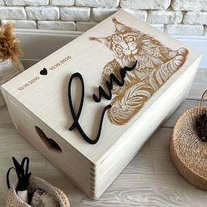 May include: A light-coloured wooden box with an engraved cat design on the lid. The lid also features the name "Luna" in black script and dates. The box has a heart-shaped cutout on the side.