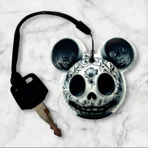 May include: A black and white key chain with a skull design featuring floral details. The skull is shaped like Mickey Mouse with large ears.