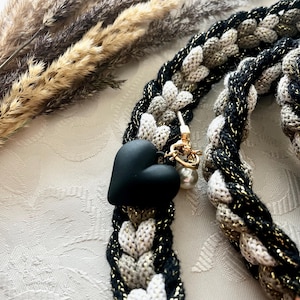 Puede incluir: Correa trenzada en negro, dorado y beige con un dije de corazón negro y herrajes dorados. La correa está hecha de un material grueso y tejido. Una pequeña cuenta blanca con forma de perla cuelga de los herrajes.