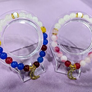 May include: Two bracelets with gold crescent moon charms. One bracelet has white, blue, red, and yellow beads. The other bracelet has white and pink beads.