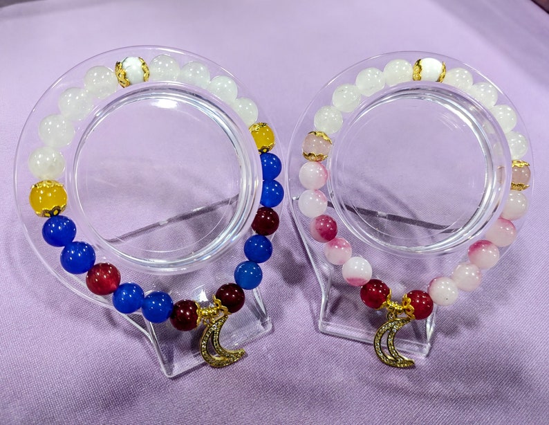 May include: Two bracelets with gold crescent moon charms. One bracelet has white, blue, red, and yellow beads. The other bracelet has white and pink beads.