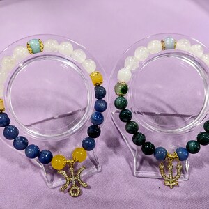 May include: Two beaded bracelets with gold charms. One bracelet has blue, white, and yellow beads with a gold trident charm. The other bracelet has green, white, and blue beads with a gold trident charm.