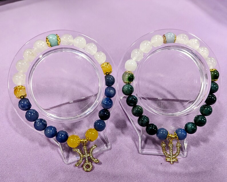 May include: Two beaded bracelets with gold charms. One bracelet has blue, white, and yellow beads with a gold trident charm. The other bracelet has green, white, and blue beads with a gold trident charm.
