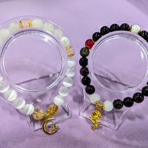 May include: Two bracelets with gold charms. One bracelet is made of white stones and has a crescent moon charm. The other bracelet is made of black and green stones and has a rose charm.