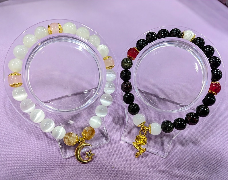 May include: Two bracelets with gold charms. One bracelet is made of white stones and has a crescent moon charm. The other bracelet is made of black and green stones and has a rose charm.